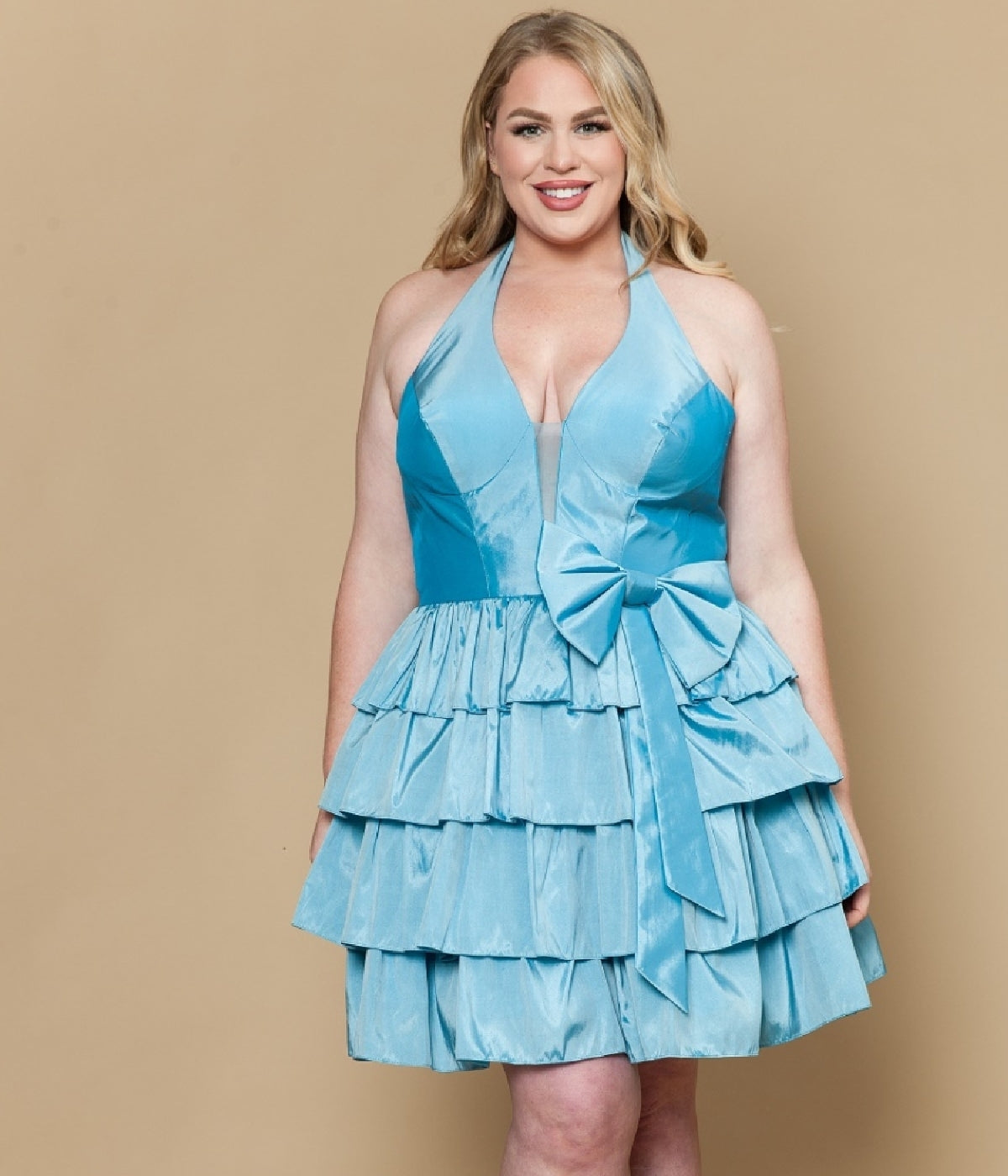 A woman models the Plus-Size Short Halter Party Dress: Poly USA W1216 in blue, featuring tiered ruffles and a large waist bow, against a plain beige background.